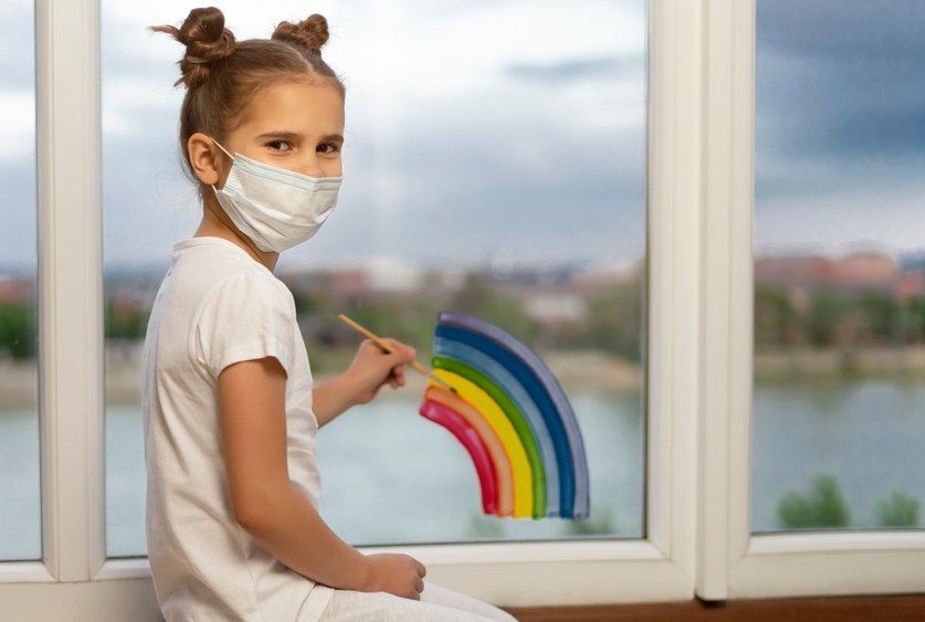 a child wearing a mask painting a rainbow on a window 