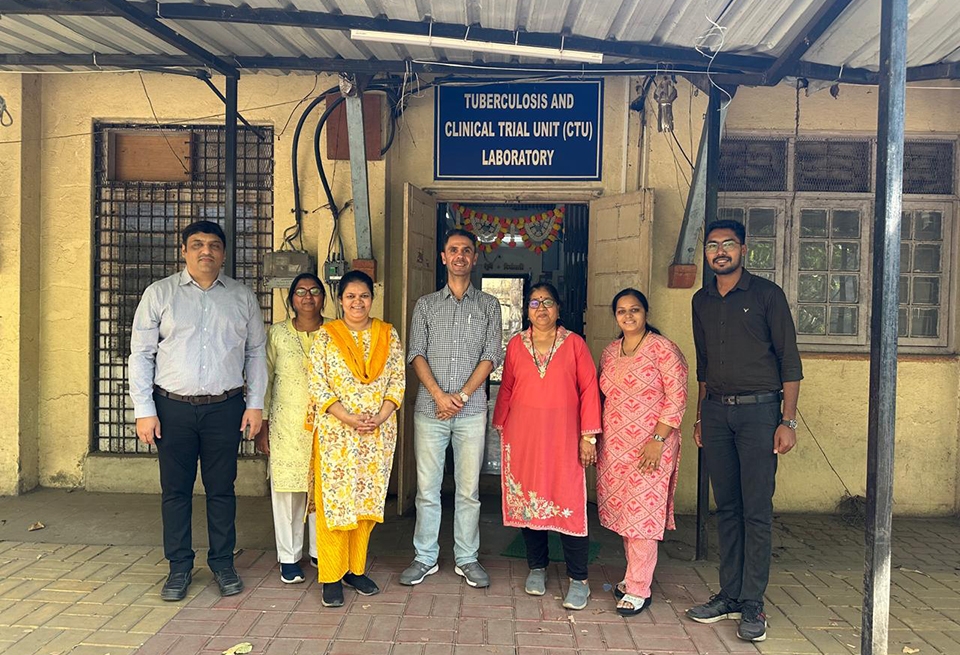 six people standing in front of a building with a sign reading "Tuberculosis and Clinical Trial Unit (CTU) Laboratory"