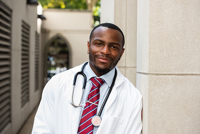 A male medical student wearing a white coat, with a stethoscope around his neck, leans against the side of a building.