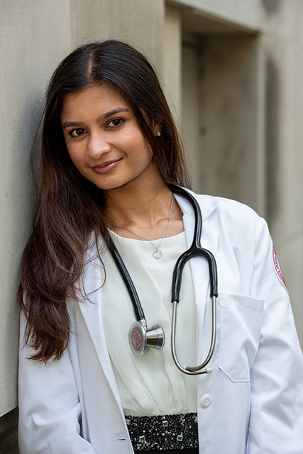 A female medical student in a white coat, with a stethoscope around her neck, leaning against the side of a building.