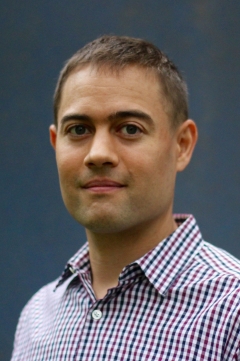 a fair-skinned man wearing a plaid shirt posing for a portrait in front of a blue background.