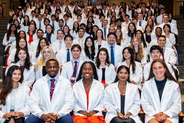 Wide shot of students seated in audience wearing white coats 
